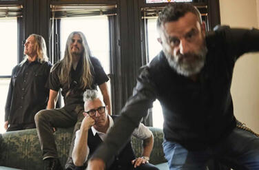 The four members of the band Tool. One seems to be moving towards the camera, another is sitting on a large couch, a third is sitting on top of the back of the couch, and the fourth is standing behind it.